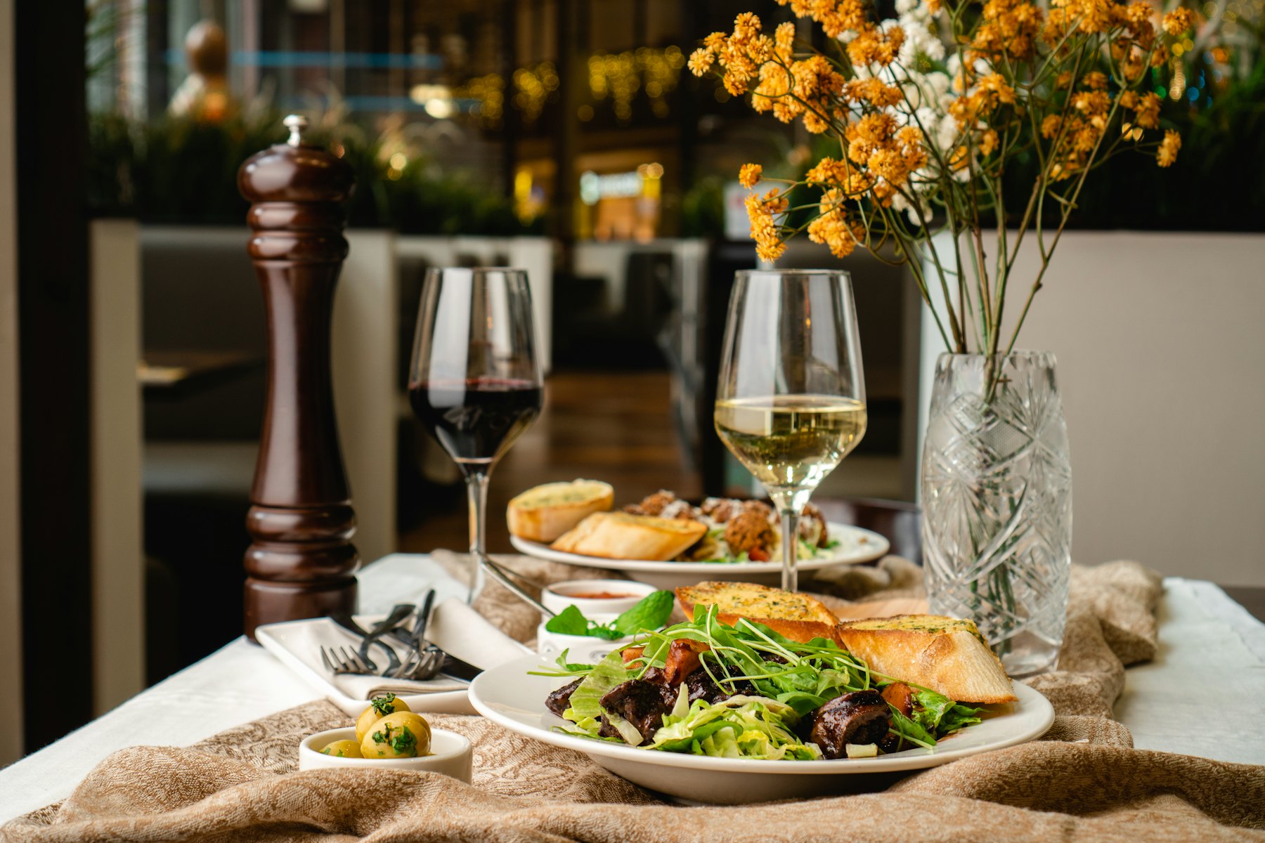 Nakryty stolik w bistro: sałatka, pieczywo, kieliszki wina i bukiet żółtych kwiatów